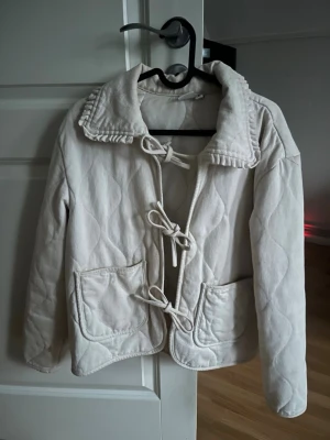 Vit/beige jacka med knyt - Superfin vårjacka med stora fickor framtill och dekorativa knytningar som stängning. Jackan har en bred krage med volangdetaljer och långärmade ärmar. Perfekt för både vår och sommar! Den är hel och i ett bra skick, men har två små fläckar som knappt syns men går bort i tvätt. Storlek M men passar även S.