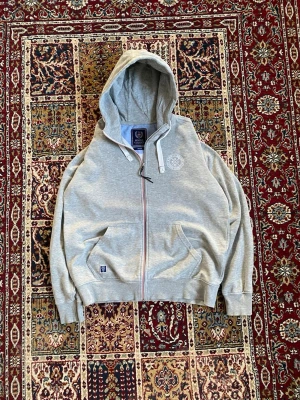 Grå hoodie från Marine Classics - Snygg grå hoodie från Marine Classics med dragkedja, huva och två fickor framtill. Broderad logga på bröstet och en liten patch vid fickan. Mjuk bomullsblandning och ribbade muddar vid ärmslut och midja. Perfekt för en avslappnad stil.