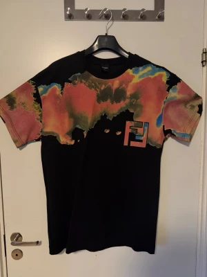 Färgexplosiv t-shirt från Fendi - Unik svart t-shirt från Fendi med färgstarkt abstrakt mönster i rött, gult, blått och grönt över axlar och ärmar. Stor Fendi-logga på bröstet. Skön passform och tillverkad i mjuk bomull. Perfekt för dig som vill sticka ut.    !!!!!!!!!! Oanvänd !!!!!!!!!