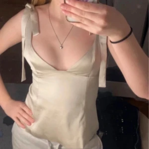 Champagnefärgad linnetopp med knyt - Snygg champagnefärgad linnetopp med knyt på axlarna och djup v-ringning. Materialet är silkesliknande och ger en lyxig känsla. Perfekt för dig som vill ha en enkel men elegant topp med fina detaljer. Tyvärr en liten fläck men den går bort bara man tvättar.