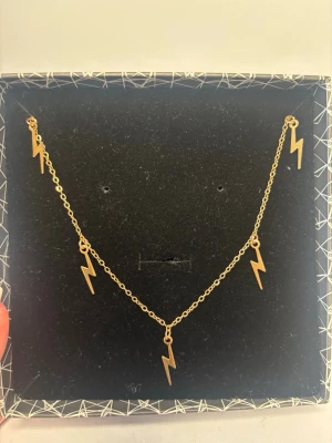 Guldigt halsband med blixtberlocker - Snyggt halsband i guldfärgad metall med fem blixtformade berlocker. Kedjan är tunn och elegant, perfekt för att ge en cool touch till din stil. Berlockerna är jämnt fördelade längs kedjan och har en modern, edgy design.
