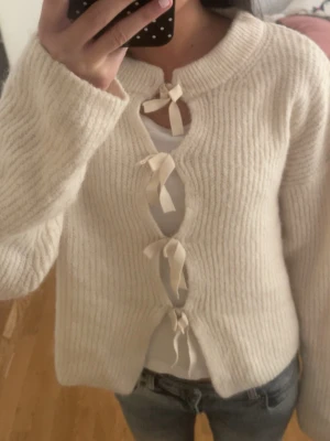 Stickad tröja med rosett - Snygg beige stickad tröja med långa ärmar och unika rosett-detaljer framtill. Tröjan har en rak, något kortare passform och ribbstickade muddar. Perfekt för dig som gillar en söt och trendig look.