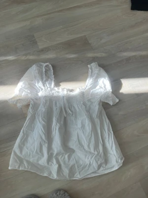 Vit puffärm blus med smock - Söt vit blus med puffärmar och smockad överdel. Blusen har en rundad halsringning med knytband och är gjord i ett lätt och luftigt material, perfekt för en trendig och avslappnad look. Passar dig som gillar romantiska detaljer och stilrena plagg.