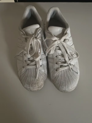 Vita Adidas Superstar sneakers - Klassiska vita Adidas Superstar sneakers med ikoniska tre ränder på sidorna och snäckformad tå. Skorna har vita skosnören och är tillverkade i läder med detaljer i gummi.