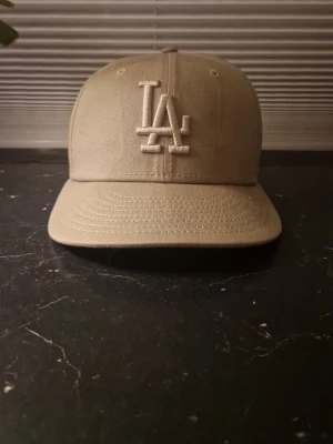 Beige LA-keps från New Era - Snygg beige keps från New Era med LA-logga broderad framtill. Klassisk modell med böjd skärm och ventilerande hål upptill. Tillverkad i slitstark bomull och har New Era-loggan på sidan. Perfekt för dig som gillar streetwear och sportig stil.