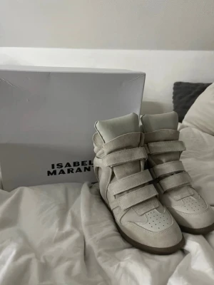 Isabel Marant beige höga sneakers - Snygga höga sneakers från Isabel Marant i beige mocka och läder. Ny pris 6500kr!!! Skorna har tre breda kardborreband, vadderad plös och en chunky sula i gummi. Perfekta för dig som vill ha en trendig och unik look med streetkänsla.
