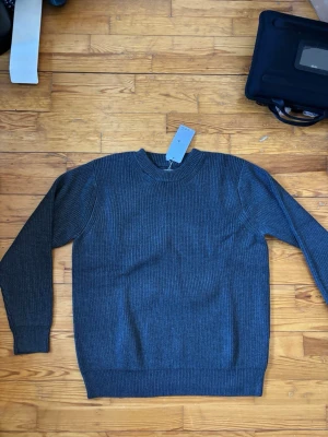 Mörkgrå stickad tröja Maison Margiela - Snygg mörkgrå stickad tröja från Maison Margiela med rund halsringning och ribbade muddar vid ärmslut och nederkant. Tröjan har en klassisk, rak passform och är tillverkad i ett mjukt, värmande material. Perfekt för lager på lager under kyliga dagar.