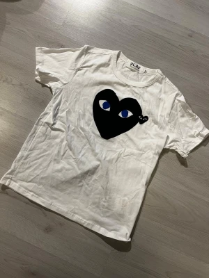 Vit Comme des Garçons PLAY t-shirt - Säljer en vit t-shirt från Comme des Garçons PLAY med det ikoniska svarta hjärtat med blå ögon tryckt på bröstet. Klassisk rund hals och korta ärmar. Perfekt för dig som gillar streetwear och vill sticka ut lite extra.