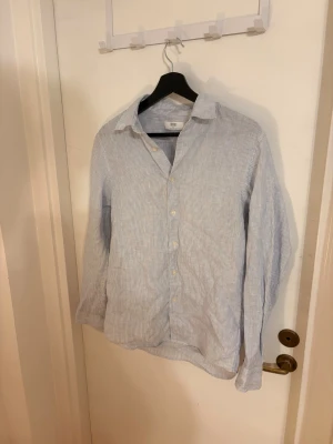 Randig skjorta från Uniqlo XS - Säljer en ljusblå och vit randig skjorta från Uniqlo i storlek XS. Skjortan har klassisk krage, knappar framtill och långa ärmar.  Perfekt för en avslappnad och stilren look. Använd 1 gång!