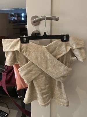 Beige offshoulder topp med volang - Trendig beige offshoulder topp med bred volangkant och omlott-detalj framtill. Mjuk och stretchig känsla, perfekt för dig som gillar en unik och snygg stil. Passar till både jeans och kjol för en cool look.