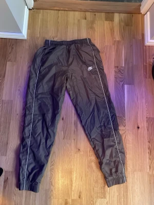 Nike svart träningsset med vita detaljer - Nike träningsset med jacka och byxor i svart med vita detaljer och logga på bröstet och benet. Setet har dragkedja på jackan, resår i midjan och vid benslut samt smala vita linjer längs ärmar och ben. Perfekt för träning eller chill. Till både man och kvinna.