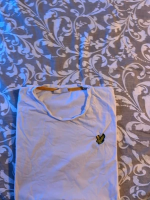 Vit t-shirt från Lyle & Scott - Klassisk vit t-shirt från Lyle & Scott med den ikoniska gula örnloggan broderad på bröstet. T-shirten har rund hals och korta ärmar. Perfekt basplagg för en clean och enkel stil.