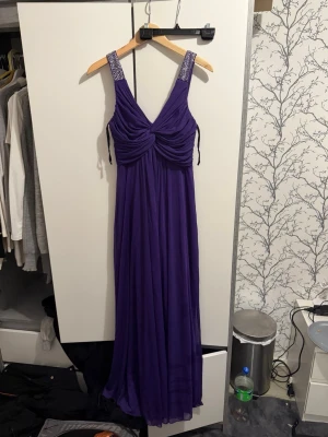 Lång lila klänning från Dollar - Säljer en elegant långklänning i lila från Dollar. Klänningen har breda axelband dekorerade med glittrande stenar och ett draperat liv med v-ringning. Tyget faller mjukt och ger en flowy siluett, perfekt för speciella tillfällen.