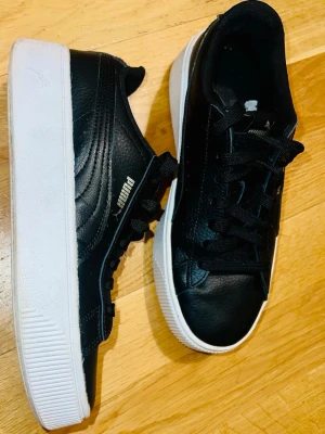 Svarta Puma sneakers med platåsula - Snygga svarta sneakers från Puma med klassisk vit platåsula och svart läderimitation. Skorna har svarta skosnören, diskret Puma-logga på sidan och tungan samt stilren design som passar till det mesta. Perfekta för dig som gillar streetstyle och vill ha en clean look.