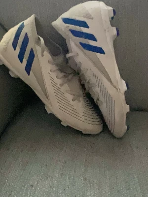 Adidas predator  - Säljer ett par vita fotbollsskor från Adidas med klassiska blå ränder på sidan. Skorna har snörning, låg profil och dobbar för bra grepp på planen. Tillverkade i syntetmaterial med stilrena svarta linjer på ovandelen. Perfekta för dig som vill sticka ut på planen.