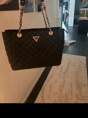 Svart  handväska från Guess - Säljer en svart quiltad handväska från Guess med guldfärgade kedjehandtag och Guess-logga i metall framtill. Väskan har en klassisk rektangulär form och är tillverkad i konstläder med rutmönster. Perfekt för dig som gillar stilrena och trendiga accessoarer.