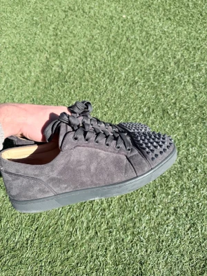 Svarta Louboutin sneakers med nitar - Säljer ett par svarta sneakers från Christian Louboutin i mocka med coola nitar på tån och klassiska röda sulor. Skorna har svarta skosnören och en låg profil som ger en clean och edgy look.