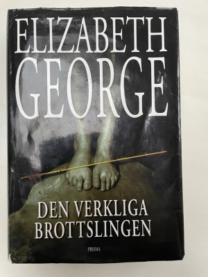 Den verkliga brottslingen - En spännande kriminalroman av Elizabeth George med svart hård pärm och omslag som visar fötter och en pil. Boken är på svenska och handlar om deckarparet Thomas Lynley och Barbara Havers. Perfekt för dig som gillar välskrivna deckare.