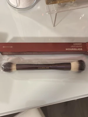 Hourglass Ambient dubbelsidig sminkborste - Säljer en lyxig dubbelsidig sminkborste från Hourglass, Ambient. Borsten har ett elegant brunt handtag med guldfärgad logga och mjuka, täta syntetiska borst i vitt med mörka toppar. Perfekt för applicering av puder, highlighter eller bronzer.