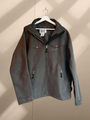 Grå vindjacka från Jack Wolfskin - Vintage, Mörkgrå vindjacka från Jack Wolfskin med stormlock, blixtlås och dragsko. Luva som kan döljas inuti kragen. Bröstfickor med knappar och sidofickor med dragkedja. Mkt fint skick.