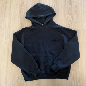 Svart hoodie Essentials Fear of God - Svart oversized hoodie från Essentials Fear of God med stor huva och känguruficka. Diskret logga på bröstet och stor 'ESSENTIALS FEAR OF GOD'-text på ryggen. Tillverkad i mjukt bomullsmaterial, perfekt för en avslappnad streetwear-look.