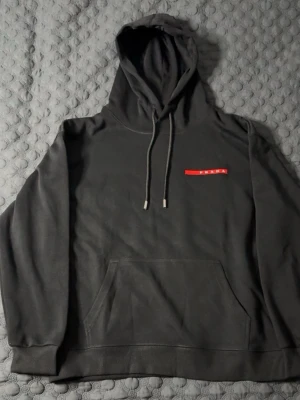 Svart hoodie från Prada - Snygg svart hoodie från Prada med klassisk röd logga på bröstet. Hoodien har dragsko i huvan och en stor magficka framtill. Tillverkad i mjukt bomullsmaterial som känns skönt mot huden. Perfekt för dig som gillar stilrena designer och exklusiva märken. STORLEK M, PASSAR M OCH S. HELT NY, PRISLAPP PÅ!!