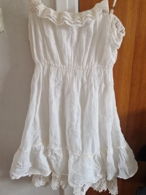 Vit kortklänning med spetsdetaljer - Söt vit kortklänning med volangkant och spetsdetaljer vid halsringning och nederkant. Klänningen har en snygg passform och är ärmlös, perfekt för varma dagar. Tillverkad i ett lätt bomullstyg som ger en luftig känsla. Något genomskinlig och behövs stycka