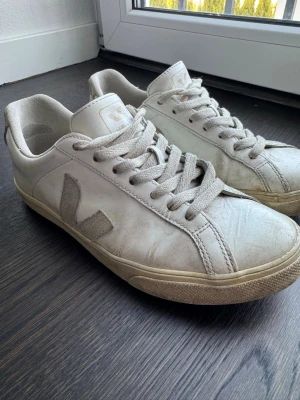 Vita Veja sneakers - Säljer ett par klassiska vita Veja sneakers med den ikoniska V-loggan i mocka på sidan. Skorna har vita snören, låg profil och en beige sula. Perfekta för dig som gillar stilrena och tidlösa sneakers.