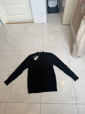 Svart stickad tröja Polo Ralph Lauren - Klassisk svart stickad tröja från Polo Ralph Lauren med rund halsringning och diskret röd logga broderad på bröstet. Tröjan har långa ärmar och ribbade muddar. Perfekt för en stilren och enkel look.