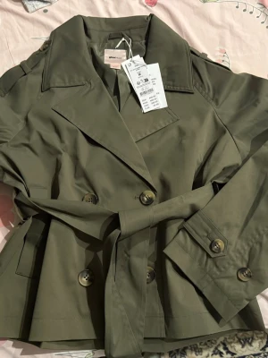 Kort khakigrön trenchcoat med bälte - Stilren kort trenchcoat från Gina Tricot i khakigrönt bomullstyg. Jackan har bred krage, dubbelknäppning framtill, axelklaffar och ett matchande knytbälte i midjan. Ärmarna har dekorativa knappar vid mudden och jackan har en klassisk stormflik bak.