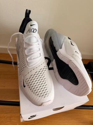 Nike Air Max 270  - Nike Air Max 270 sneakers i vitt med svarta detaljer och stor synlig Air-enhet i hälen. Ovandelen är i mesh för bra ventilation och har en modern, sportig look. Klassisk Nike-logga på sidan och tungan. Perfekta för dig som gillar streetstyle och komfort.