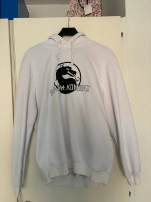 Vit Mortal Kombat hoodie med tryck - Snygg vit hoodie med svart Mortal Kombat-logga och dragsko i huvan. Hoodien har en klassisk passform och är perfekt för dig som gillar gaming och streetwear. Mjuk bomullskänsla och ribbade muddar vid ärmslut och nederkant.