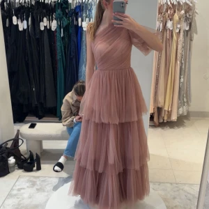 Lång rosa tyllklänning oneshoulder - Romantisk långklänning i rosa tyll med flera lager och oneshoulder-design. Klänningen har volanger och markerad midja, perfekt för speciella tillfällen. Den luftiga tyllen ger en drömmig känsla och den asymmetriska axeln gör looken unik.