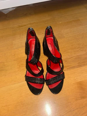 Svarta pumps från Cesare Paciotti - Snygga svarta pumps från Cesare Paciotti med flera remmar i ormmönstrat läder och röd innersula i skinn. Skorna har öppen tå och dragkedja bak för enkel påtagning. Perfekta för dig som vill sticka ut med en unik design.