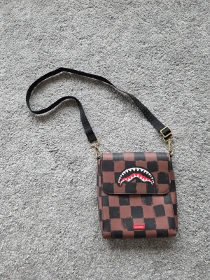 Brun axelväska Sprayground - Cool brun axelväska från Sprayground med svart rutmönster och hajtandsmotiv på locket. Väskan har justerbar svart axelrem och detaljer i guld. Materialet är slitstark canvas och designen är riktigt street och unik.