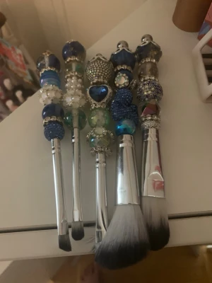 Sminkborstar med blå dekorationer - Säljer ett set med fem sminkborstar med handtag dekorerade med blå, silver och vita pärlor och stenar. Borsthuvudena är vita och svarta med mjuka syntetborst. Perfekta för dig som vill ha unika och snygga sminkverktyg i sminkväskan. Helt nytt skick.