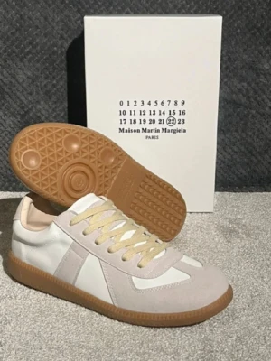 Maison Margiela  - Maison Margiela sneakers med beige mockadetaljer, vit skinnpanel och ljusgula snören. Sulan är i gummi med klassiskt mönster. Låg modell med rund tå och platt sula, perfekt för dig som gillar stilrena och lyxiga sneakers. Dom är 43 men passar 42 och innersula är inte som den vanliga men dom har ett hål i sulan på insidan av höger foten (kan skicka bild ifall det behövs).     SKOLÅDAN FÅR MAN EJ MED