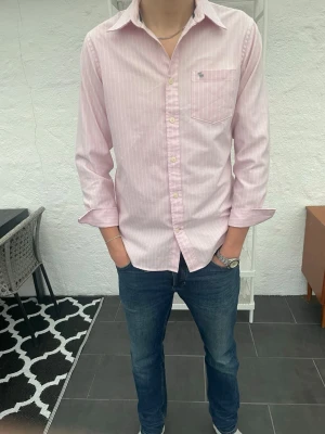 Ljusrosa randig skjorta från abercrombie and fitch - Säljer en ljusrosa skjorta från abercrombie and fitch med diskreta vita ränder. Skjortan har klassisk krage, bröstficka med broderad logga och långa ärmar med knappar vid manschetterna. Perfekt för en clean och stilren look.