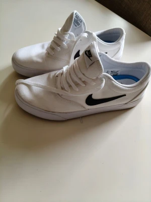 Vita Nike sneakers med svart swoosh - Snygga vita sneakers från Nike med klassisk svart swoosh på sidan. Skorna har lågt skaft, vita snören och ovandel i textil. Perfekta för en clean och sportig stil. Sulan är vit och designen är enkel men stilren.