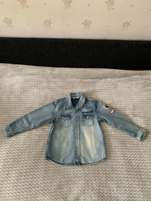 Ljusblå jeansjacka från Kids by Lindex - Snygg ljusblå jeansjacka med två bröstfickor, tryckknappar och broderad text 'NORTHAMPTON' på bröstet. Jackan har en patch på ena ärmen och klassisk krage. Perfekt för en cool och avslappnad stil.