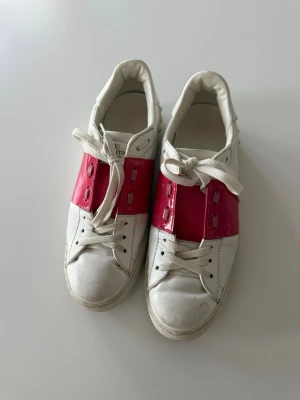 Valentino Garavani vita och rosa sneakers - Snygga sneakers från Valentino Garavani i vitt läder med en bred rosa panel över snörningen. Klassisk låg modell med rund tå och vita skosnören. Perfekta för dig som vill ha en statement-sneaker med exklusiv känsla och cool färgkombination.