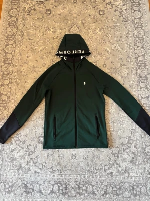 Peak Performance Hoodie stl: S - Säljer en stilren och bekväm hoodie från Peak Performance i en elegant mörkgrön färg. Perfekt som vår- och höstjacka eller som ett värmande lager under kyligare dagar.  • Storlek: S • Färg: Mörkgrön • Skick: Som ny, inga skador eller tecken på slitage • Märke: Peak Performance • Modell: Rider Essentials Zip Hood  En modern och mångsidig tröja som passar lika bra till vardags som till en aktiv livsstil.  Kan hämtas upp eller fraktas.  Hör gärna av dig vid intresse eller frågor!