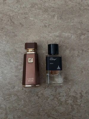 Parfym Liquid Brun & Rifaaqat Adorn  - Två parfymer: Liquid Brun i en brun och guldfärgad flaska med geometrisk design, samt Rifaaqat Adorn i en genomskinlig glasflaska med svart etikett och svart kork. Båda flaskorna har en modern och stilren look, perfekta för dig som gillar exklusiva dofter, säljer båda parfymer har använt de vid skolan och inte min typ av parfym, 90 av 100 ml återstår på båda, båda parfymer säljes! (Liquid brun lådan medkommer på köpet!)