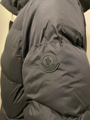Svart dunjacka från Moncler - Säljer en svart dunjacka från Moncler med huva och stora fickor framtill. Jackan har Moncler-logga på ärmen och präglad Moncler-text längs dragkedjan. Puffig modell som håller dig varm under kalla dagar. Perfekt för dig som vill ha både stil och funktion.