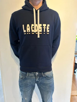 Mörkblå hoodie från Lacoste - Snygg mörkblå hoodie från Lacoste med stor vit logga framtill och vita dragsnören i huvan. Klassisk känguruficka och ribbade muddar vid ärmslut och nederkant. Perfekt för en avslappnad och trendig stil. Materialet känns mjukt och bekvämt.