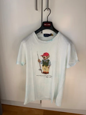 Polo Ralph Lauren: Polo Bear TieDye Limited Edition T-Shirt - TieDye t-shirt från Ralph Lauren med Polo Bear-tryck framtill. T-shirten har rund hals, korta ärmar och en slim fit passform. Tillverkad i mjuk bomull och har en cool björn med fiskespö på bröstet. Perfekt för dig som gillar streetwear och klassisk stil. 🎉Limited Edition men använd rätt ofta och har väldigt lite osynliga ljusgula små ytor under armhålorna därav pris (ordinarie pris: 1995 kronor) 🎉 För mer info skriv dm!