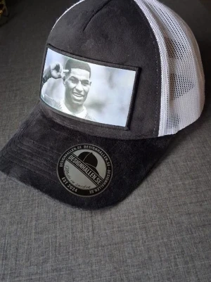 Rashford Svart och vit keps från Designhallen - Snygg svart och vit truckerkeps från Designhallen med mesh på sidorna och baksidan. Framsidan har en stor svartvit bild med motiv och böjd skärm i mockaimitation. Märke och etikett framtill. Perfekt för dig som vill sticka ut med en unik accessoar.