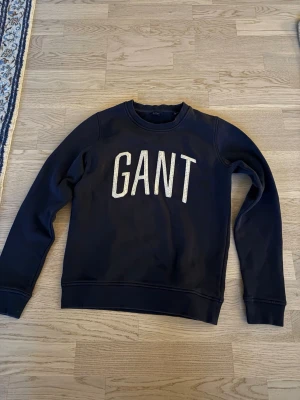 Mörkblå sweatshirt från GANT - Snygg mörkblå sweatshirt från GANT med stor vit logotyp på bröstet. Tröjan har rund halsringning, långa ärmar och ribbade muddar vid ärmslut och nederkant. Perfekt för en avslappnad och stilren look.