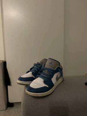 Air Jordan 1 Low blå och vita sneakers - Säljer ett par Air Jordan 1 Low sneakers i blått och vitt med grå detaljer. Skorna har klassisk låg siluett, perforerad tåbox och blå snörning. Ovandelen är i läder och textil, med den ikoniska Jumpman-loggan på plösen.