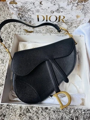 Svart Dior Saddle Bag i läder - Säljer en ikonisk svart Dior Saddle Bag i exklusivt läder med guldfärgade metalldetaljer och det klassiska D-spännet framtill. Väskan har en unik, böjd form och justerbart axelband. Perfekt statement piece för dig som vill ha något extra från Dior.